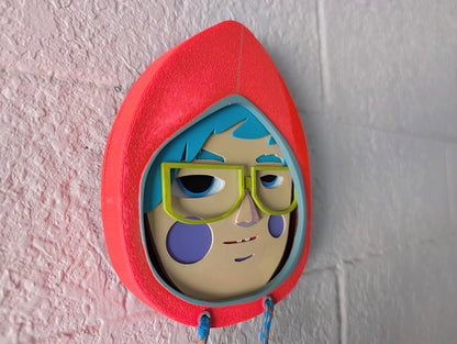 Side view showing depth of a bright orange colored hoodie shaped wall sculpture featuring teal hair, yellow green glasses, and lavender cheeks on a brick wall.