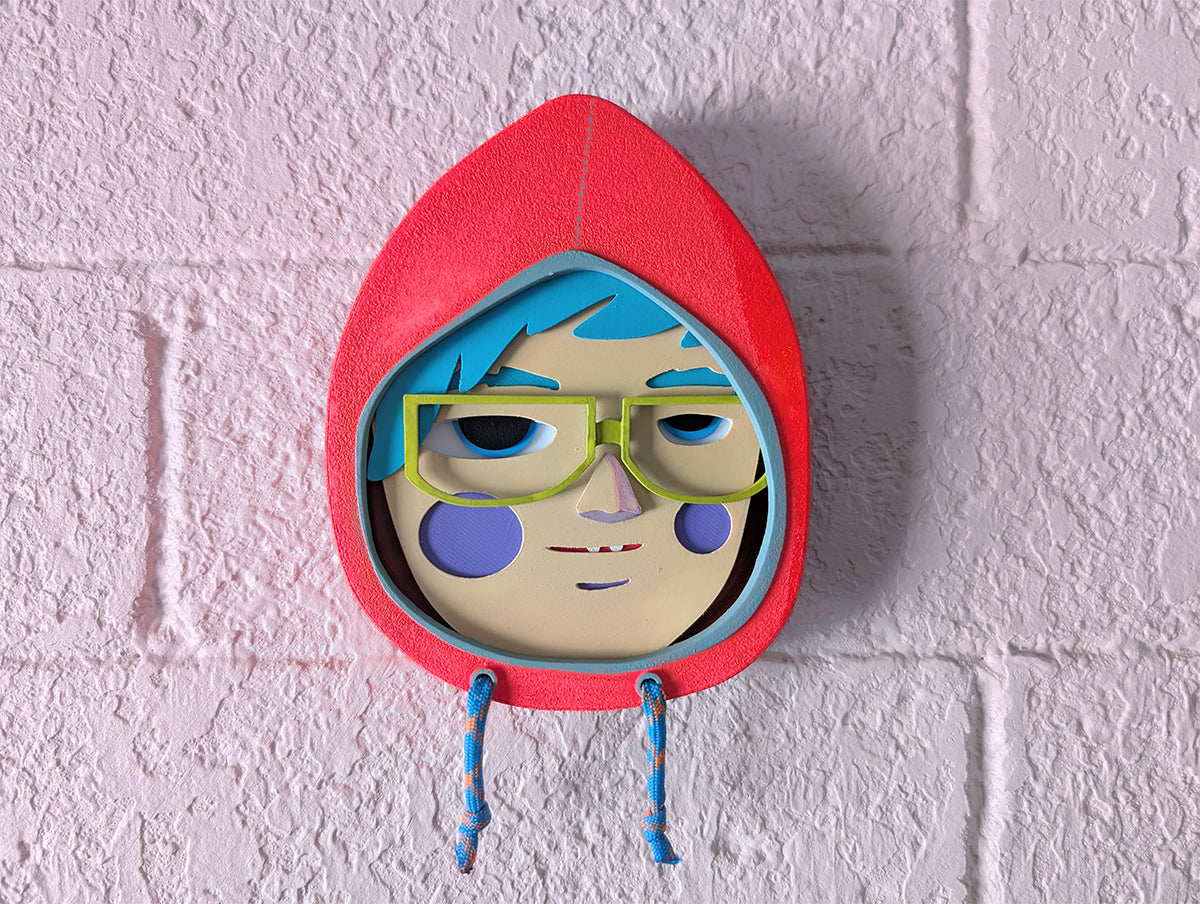 Front view of a bright orange hoodie shaped wall sculpture featuring teal hair, yellow green glasses, and purple cheeks on a brick wall.