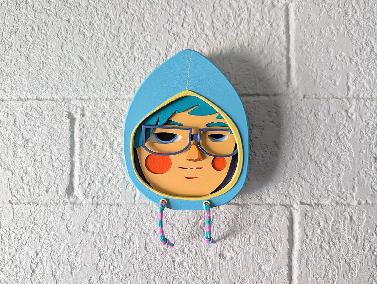 Front view of a light blue hoodie shaped wall sculpture featuring teal hair, dark blue glasses, and orange cheeks on a brick wall.