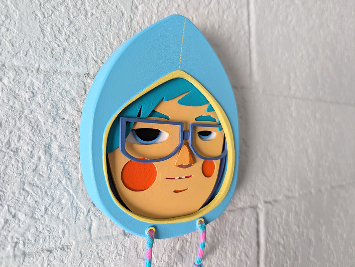 Side view showing depth of a light blue hoodie shaped wall sculpture featuring teal hair, dark blue glasses, and orange cheeks on a brick wall.
