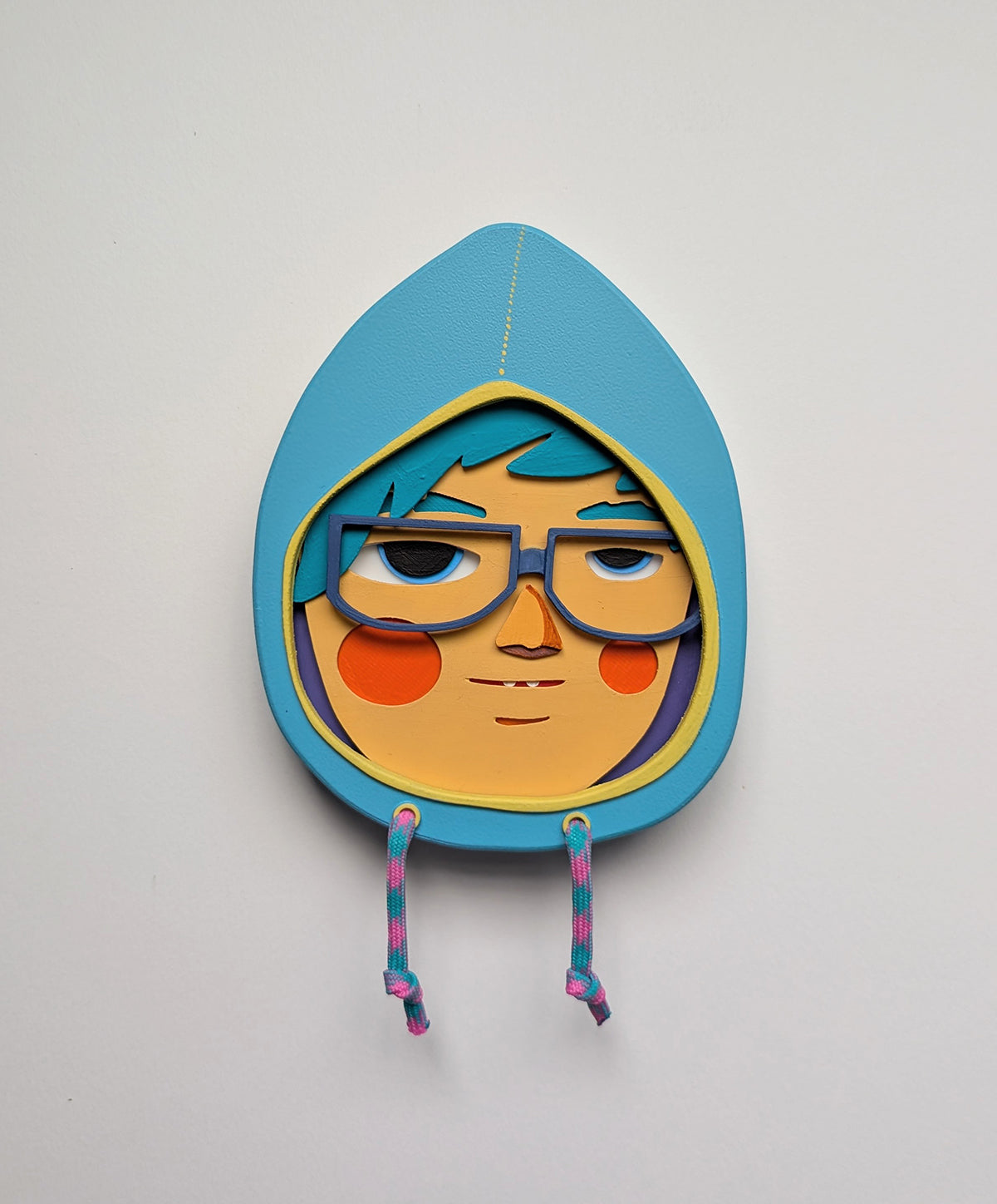 Front view of a light blue hoodie shaped wall sculpture featuring teal hair, dark blue glasses, and orange cheeks on a white background.