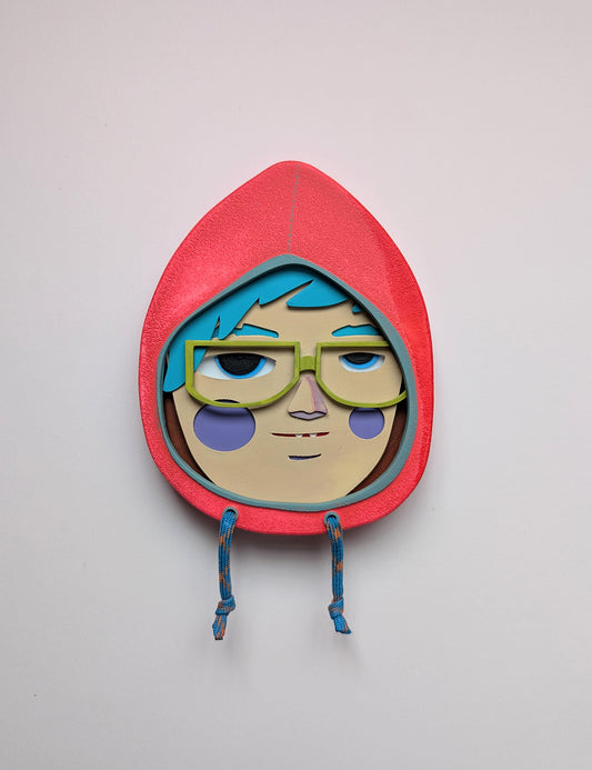 Front view of a bright orange colored hoodie shaped wall sculpture featuring teal hair, yellow green glasses, and purple cheeks on a white background.