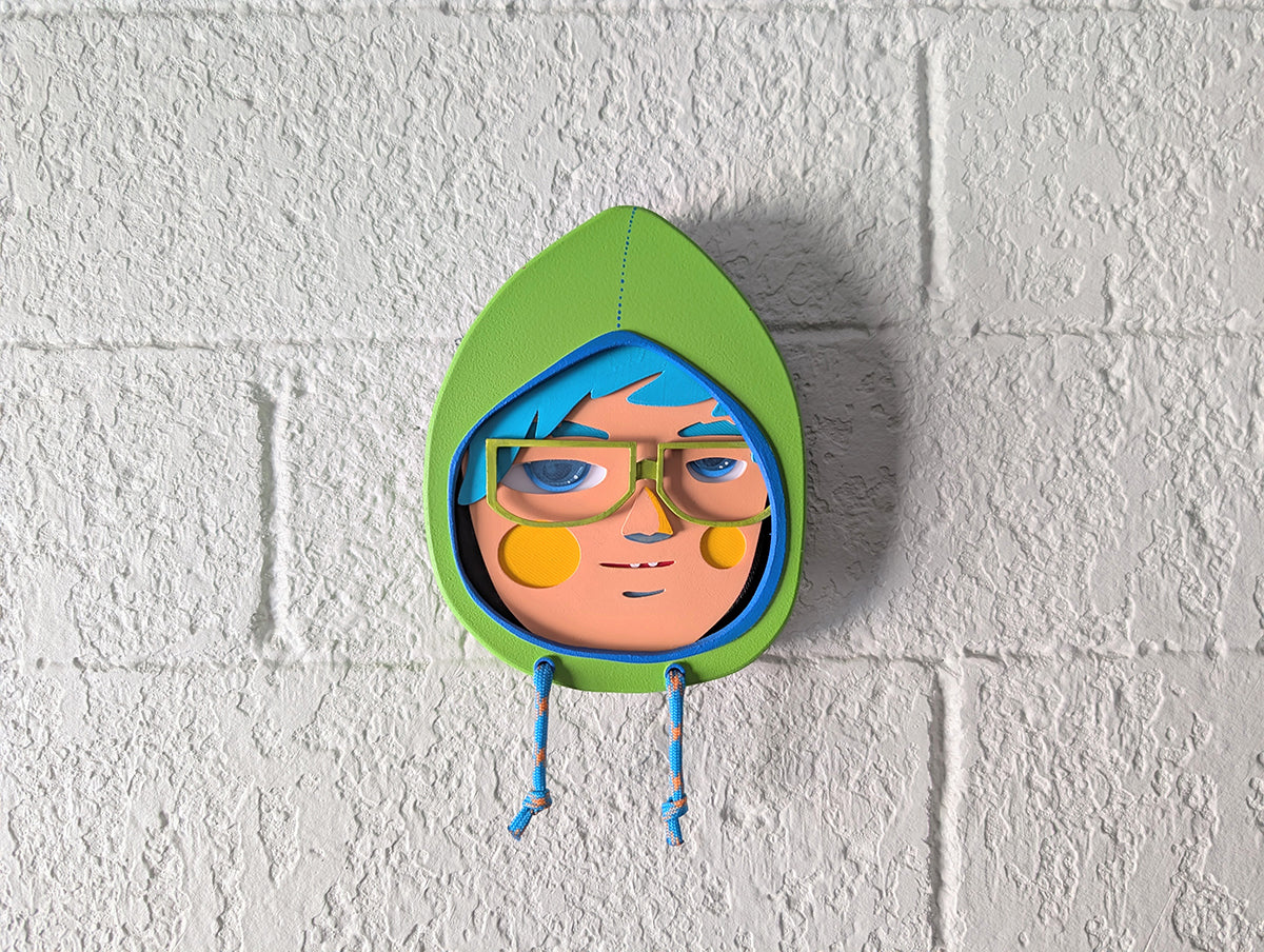 A front view showing a yellow green in color hooded wall sculpture with teal hair, green glasses hung on a brick wall.