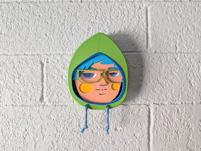 A front view showing a yellow green in color hooded wall sculpture with teal hair, green glasses hung on a brick wall.