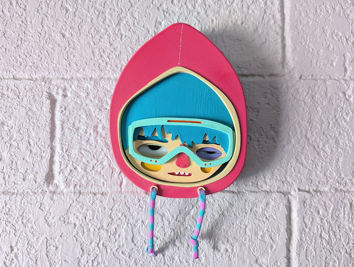 A magenta color wall sculpture featuring a character in a hoodie with teal hair, mint goggles, hung on a brick wall.
