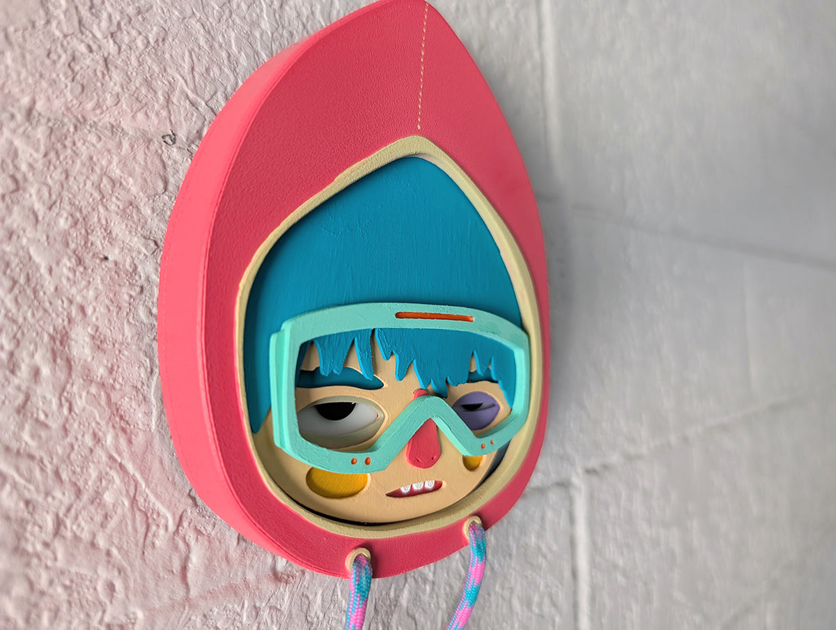 A side view showing depth of a magenta color wall sculpture featuring a character in a hoodie with teal hair, mint goggles, hung on a brick wall.