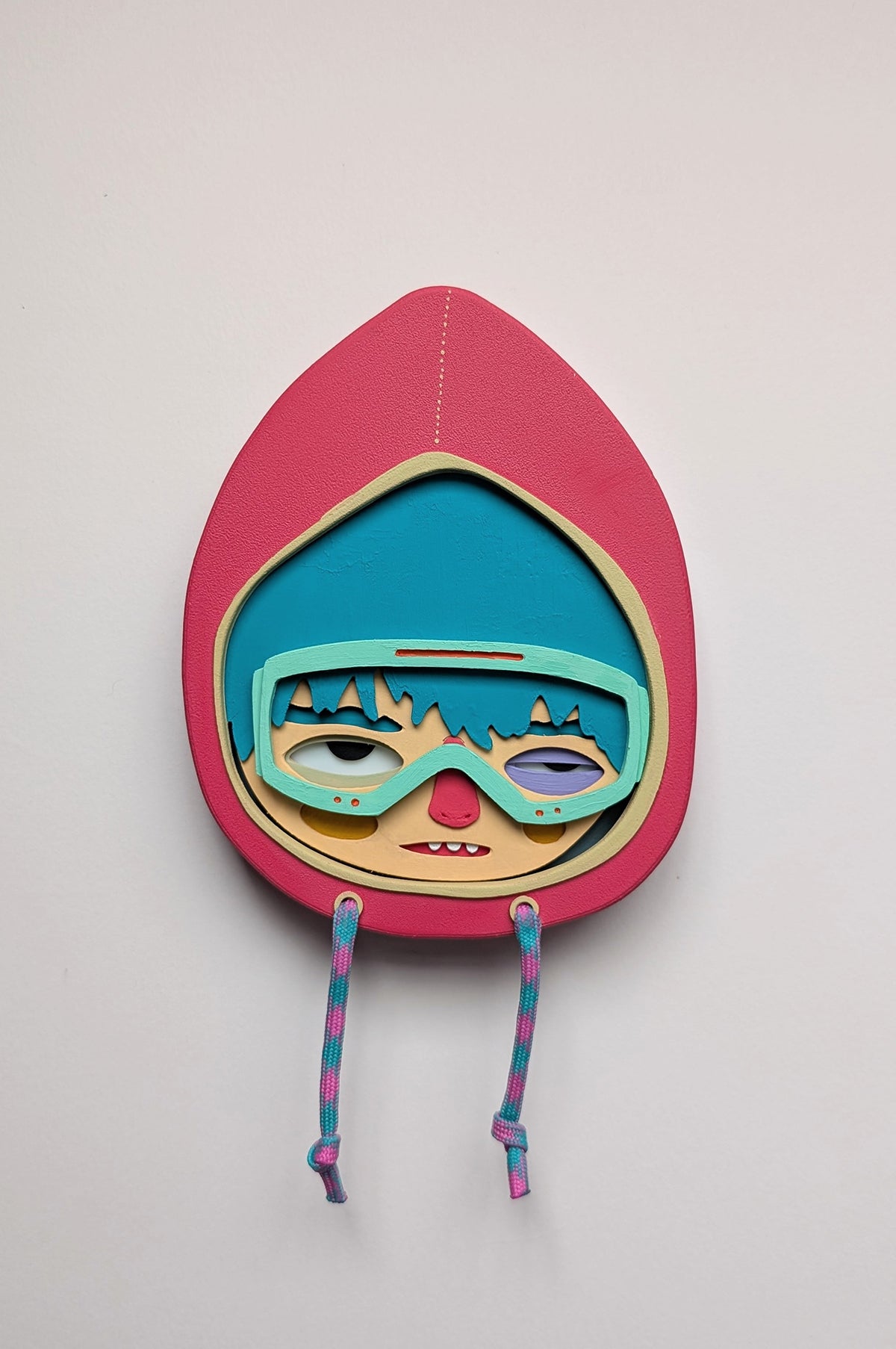 Magenta color wall sculpture featuring a character in a hoodie with teal hair, mint goggles, hung on a white wall.