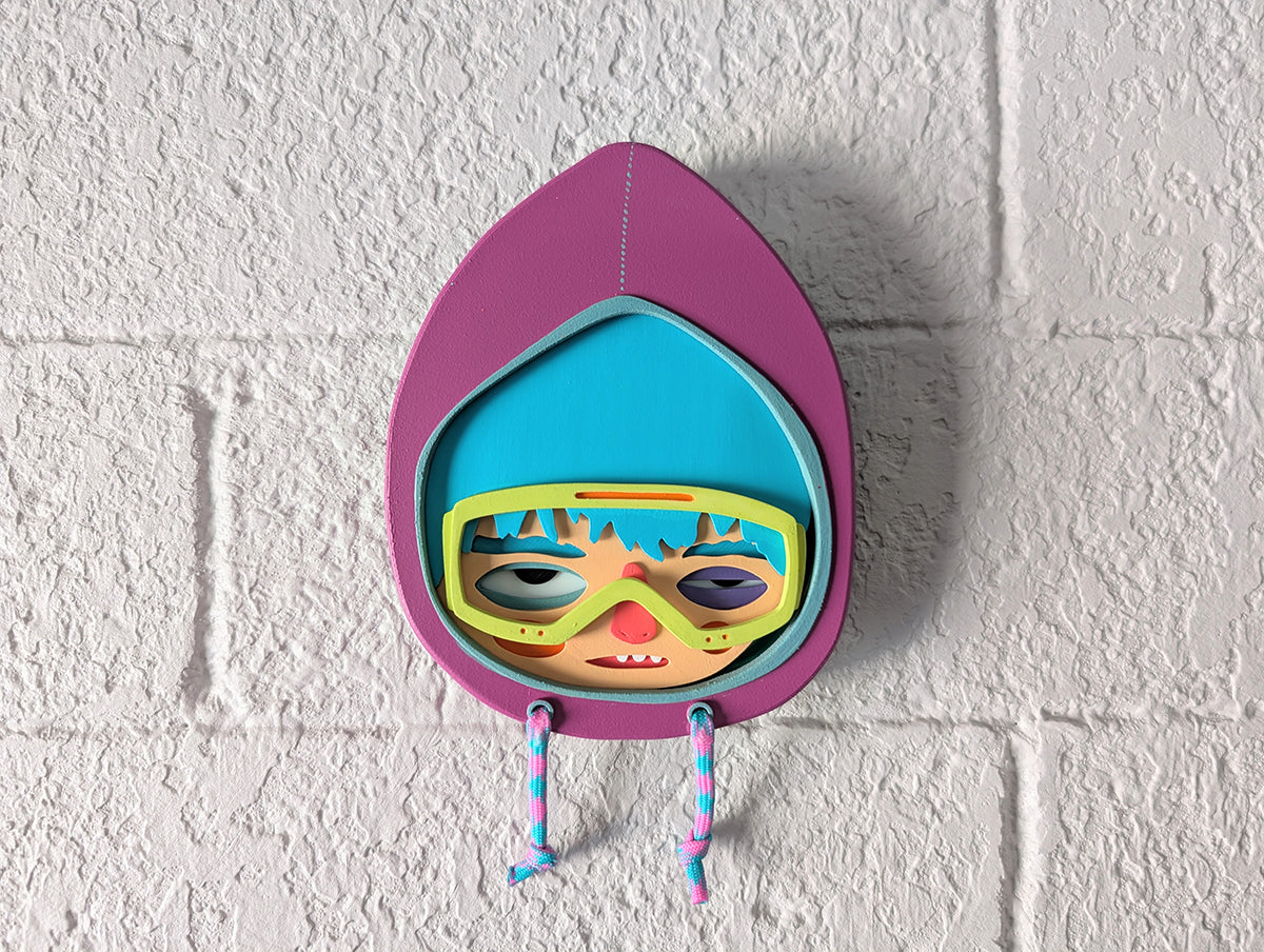 A purple hooded wall sculpture that has blue hair, bright yellow green googles on brick wall