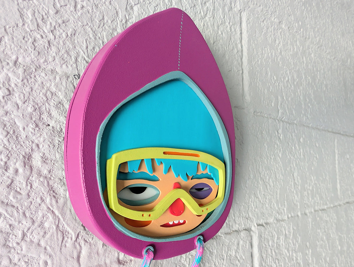 A side view of a purple hooded wall sculpture that has blue hair, bright yellow green googles on brick wall