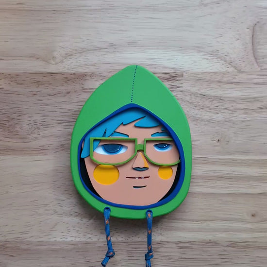 A video showing size and depth of a yellow green in color hooded wall sculpture with teal hair, green glasses with a wood table in the background.