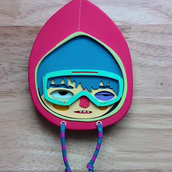 Video showing scale of a magenta colored wall sculpture featuring a character in a hoodie with teal hair, mint goggles, with a wood table in the background.