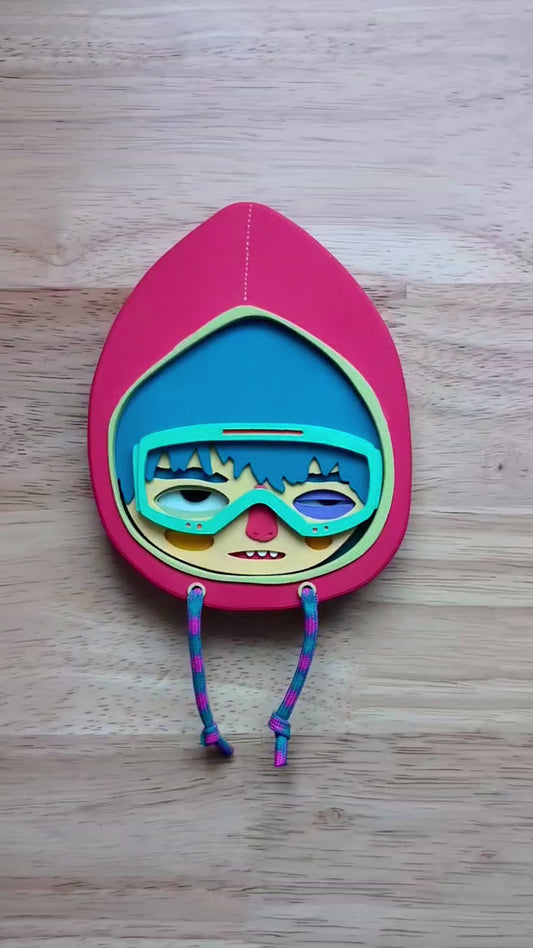 Video showing scale of a magenta colored wall sculpture featuring a character in a hoodie with teal hair, mint goggles, with a wood table in the background.